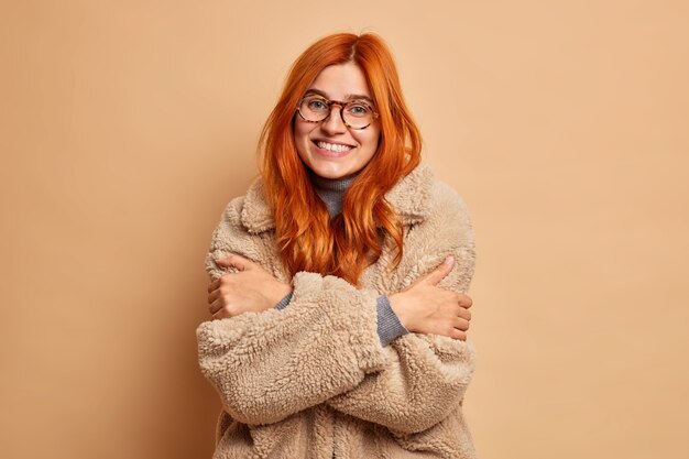 Young happy ginger young Caucasian woman dressed in cozy fur coat embraces herself to feel warm enjoys her winter holidays.