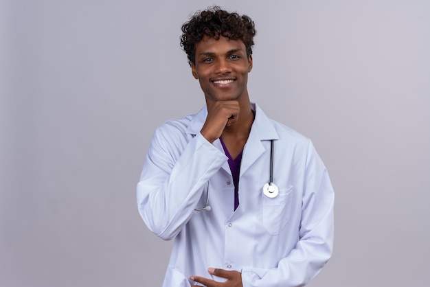 A young happy handsome dark-skinned male doctor with curly hair wearing white coat with stethoscope keeping hand on chin 