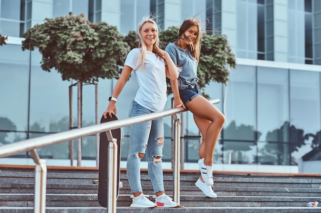 Young happy hipster girls standing on steps with a skateboard on a background of the skyscraper.