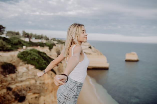 Young happy lady is sitting on a high and raising her arms up with panorama view on ocean coastline