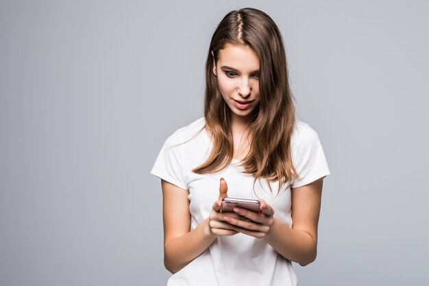 白いtシャツとブルージーンズの若い幸せな女性は白いスタジオの背景の前で携帯電話と一緒に滞在します。