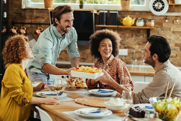 家で友達と昼食をとりながらテーブルに食べ物を持って来る若い幸せな男。