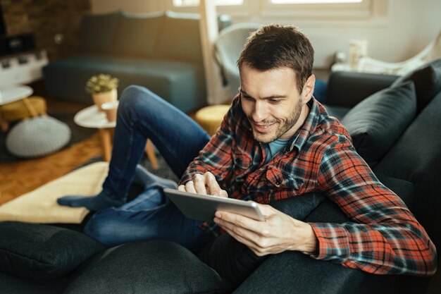 リビングルームのソファでリラックスしながらタッチパッドでインターネットを閲覧している若い幸せな男。