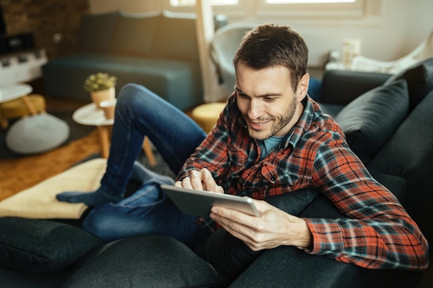 Young happy man browsing the Internet on touchpad while relaxing on the sofa in the living room.