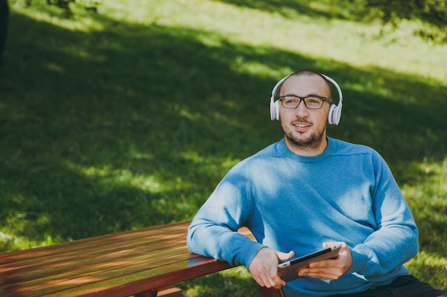 若い​幸せな​男​、​ビジネスマン​、​または​学生​が​ヘッド​フォン​で​テーブル​に​座っている​カジュアルな​青い​シャツ​の​メガネ​、​都市​公園​で​タブレット​pc​、​音楽​を​聴き​、​緑​の​自然​で​屋外​で​休憩します​。​ライフスタイル​の​レジャー​の​概念​。