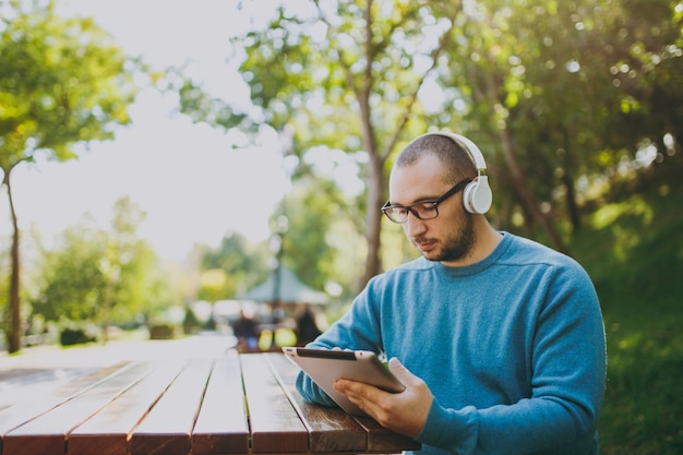 Young happy man, businessman or student in casual blue shirt glasses sitting at table with headphones, tablet pc in city park, listen music, rest outdoors on green nature. lifestyle leisure concept.