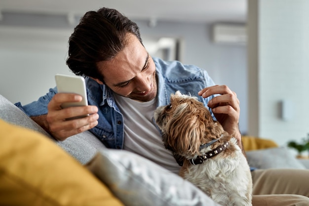 Молодой счастливый человек пользуется мобильным телефоном и развлекается со своей собакой в гостиной