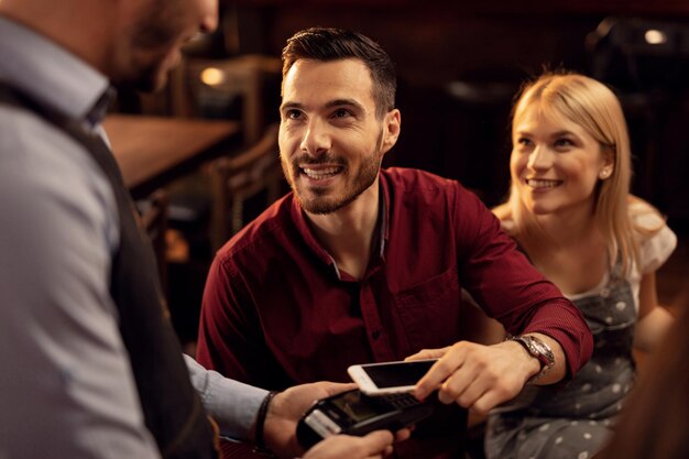 Young happy man using mobile phone while making contactless check payment in a pub