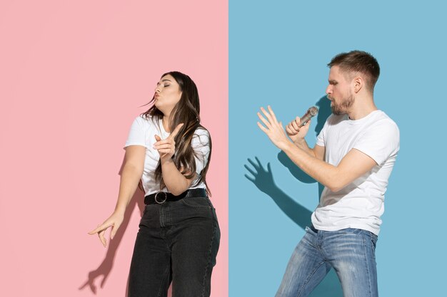 Young and happy man and woman in casual clothes on pink, blue bicolored wall, singing and dancing