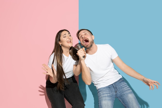 Young and happy man and woman in casual clothes on pink, blue bicolored wall, singing