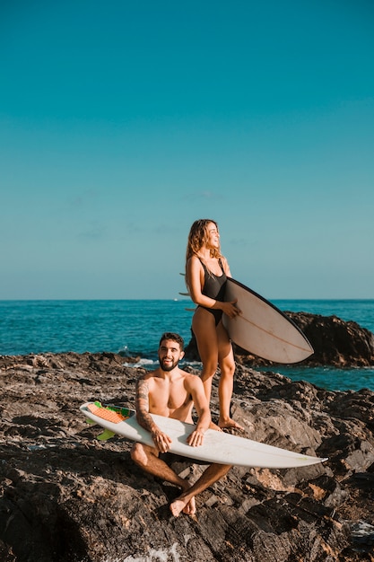 Giovane uomo felice e donna con tavole da surf sulla roccia vicino al mare