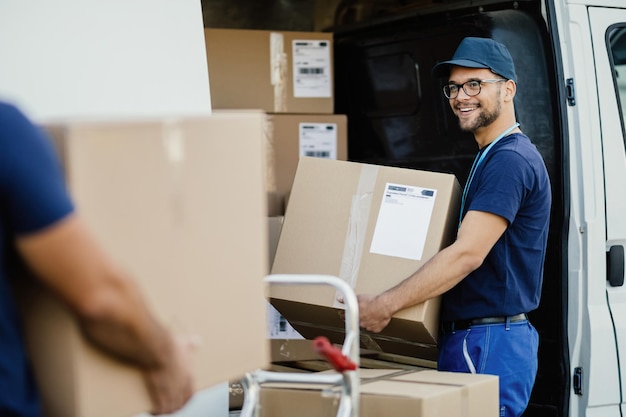 Молодой счастливый рабочий, несущий картонные коробки в фургоне доставки во время общения со своими коллегами