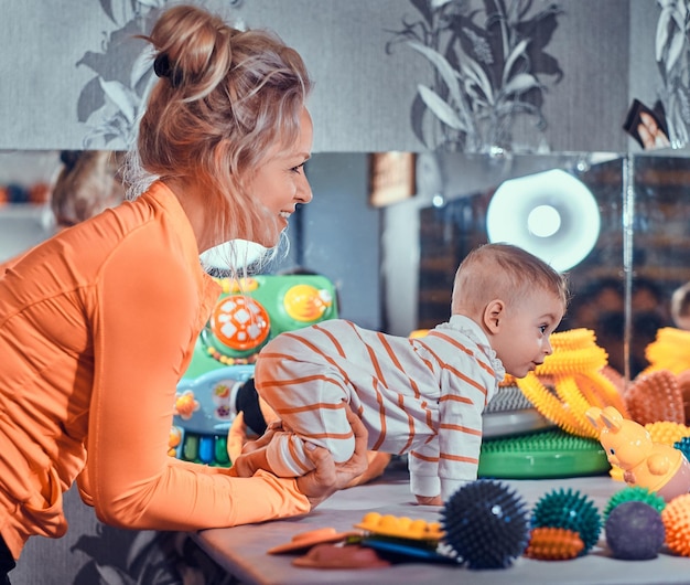 若い幸せなお母さんは、特別な治療のおもちゃでいっぱいのマッサージ師のキャビネットで、生まれたばかりの赤ちゃんと一緒に遊んでいます。