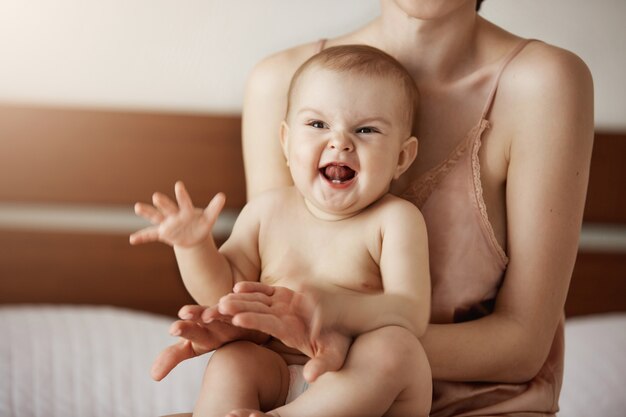 Young happy mother in sleepwear and her newborn baby sitting on bed in morning smiling playing together.