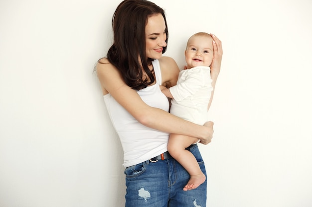 Молодая счастливая мать усмехаясь держащ смотрящ ее дочь младенца над белой стеной.