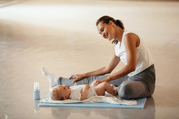 Young happy mother stretching her legs and having fun with her baby boy while working out in health club