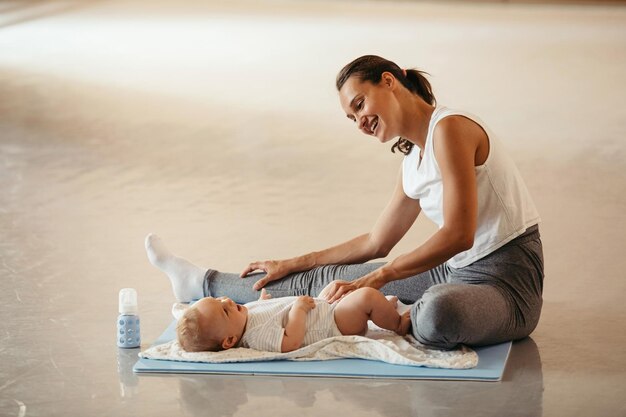 젊은 행복한 어머니는 다리를 쭉 뻗고 헬스 클럽에서 운동하는 동안 아기와 함께 즐거운 시간을 보낸다