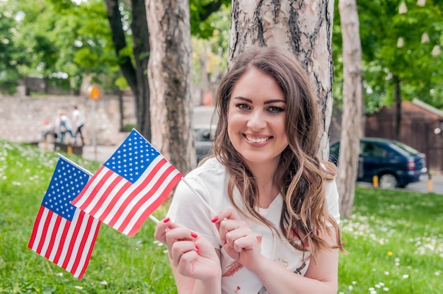 若い幸せな愛国者の女の子は、7月4日にアメリカの旗を持っています。国、愛国心、独立した日、人々の概念 - 幸せ笑顔の若い女性が白いドレスで全米の旗