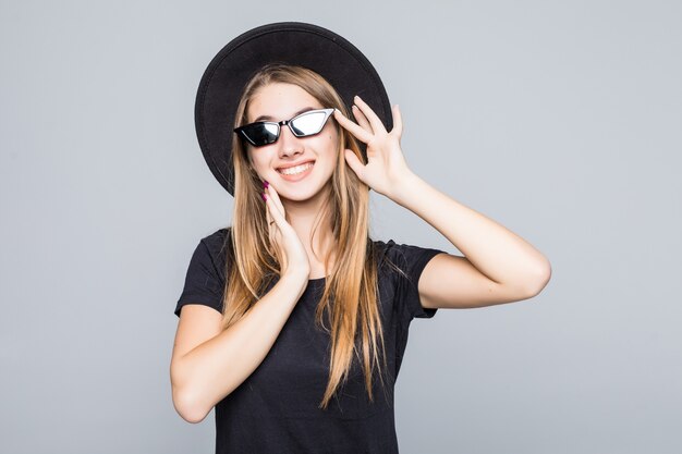Young happy pretty smiling lady in brilliant sunglasses dressed up in black hat, black t-shirt and dark pants isolated on gray background