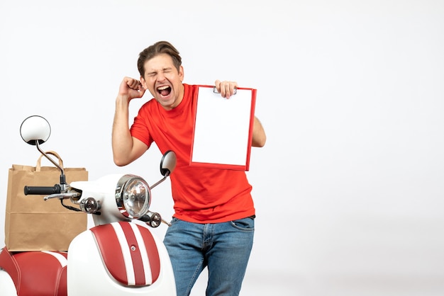 Young happy proud delivery guy in red uniform standing near scooter showing document on white wall