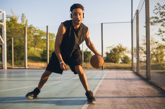 Молодой счастливый улыбающийся темнокожий мужчина занимается спортом, играет в баскетбол на рассвете, слушает музыку в наушниках, активный образ жизни, летнее утро