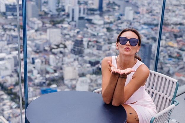 Young happy smiling caucasian woman traveller in fitting dress and sunglasses on high floor in bangkok Stylish female exploring amazing big city view