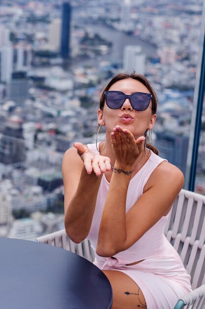 バンコクの高層階にドレスとサングラスを合わせた若い幸せな笑顔の白人女性旅行者素晴らしい大都会の景色を探索するスタイリッシュな女性