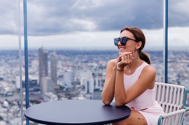 Foto gratuita viaggiatore di giovane donna caucasica sorridente felice in abito di montaggio e occhiali da sole al piano alto a bangkok elegante donna che esplora incredibile vista sulla grande città