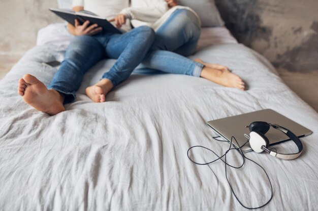 Young happy smiling couple sitting on bed at home in casual outfit reading book wearing jeans, man and woman legs romantic mood, laptop