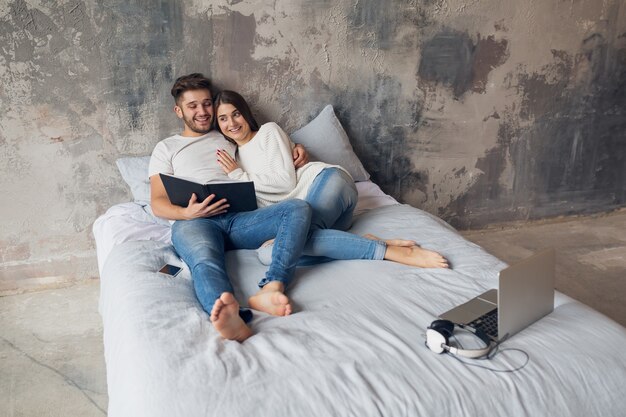 Young happy smiling couple sitting on bed at home in casual outfit reading book wearing jeans, man and woman spending romantic time together