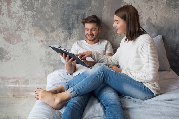 Young happy smiling couple sitting on bed at home in casual outfit reading book wearing jeans, man and woman spending romantic time together