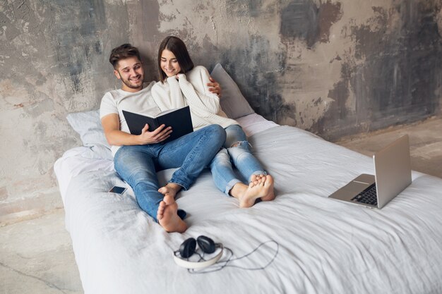 Young happy smiling couple sitting on bed at home in casual outfit reading book wearing jeans, man and woman spending romantic time together