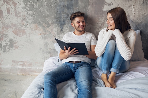 Young happy smiling couple sitting on bed at home in casual outfit reading book wearing jeans, man and woman spending romantic time together