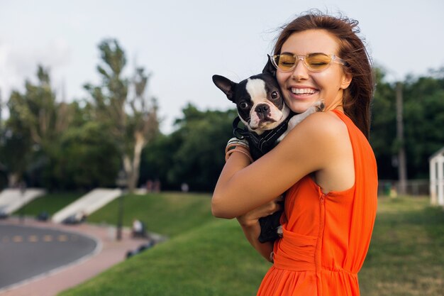 公園でボストンテリア犬を保持している若い幸せな笑顔の女性、夏の晴れた日、陽気な気分、ペットと遊ぶ、抱擁、オレンジ色のドレス、サングラス、夏のスタイルを身に着けている