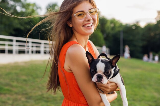 Young happy smiling woman holding boston terrrier dog in park, summer sunny day, cheerful mood, playing with pet, waving long hair, having fun, summer fashion trend