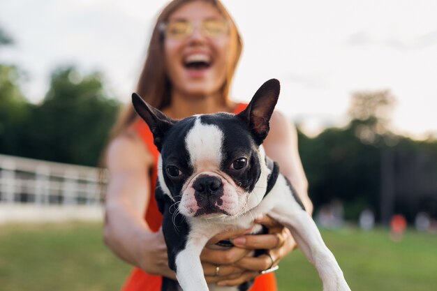 Young happy smiling woman holding boston terrrier dog in park, summer sunny day, cheerful mood, playing with pet, waving long hair, having fun, summer fashion trend