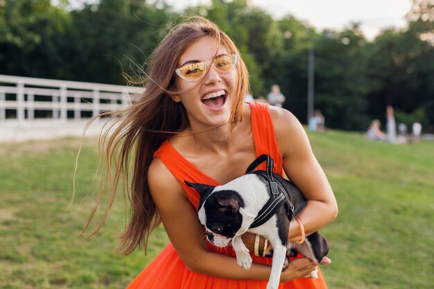公園でボストンテリア犬を保持している若い幸せな笑顔の女性、夏の晴れた日、陽気な気分、ペットと遊ぶ、長い髪を振る、楽しんで、夏のファッショントレンド
