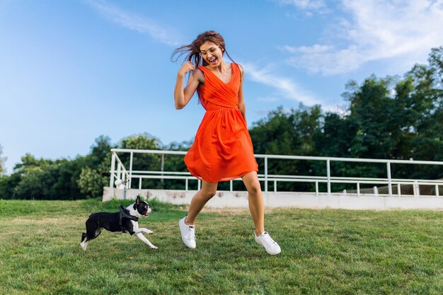 公園、夏のスタイル、陽気な気分で犬と一緒に走って楽しんでオレンジ色のドレスの若い幸せな笑顔の女性