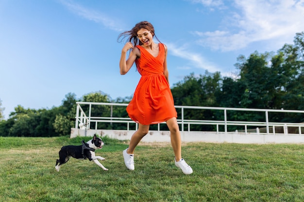 Young happy smiling woman in orange dress having fun playing running with dog in park, summer style, cheerful mood