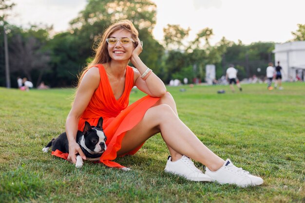 Young happy smiling woman in orange dress having fun playing with dog in park, summer style, cheerful mood