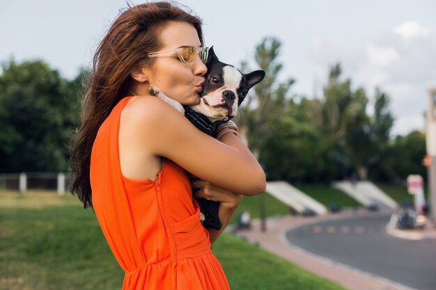 Молодая счастливая улыбающаяся женщина в оранжевом платье с удовольствием играет с собакой в парке, летний стиль, веселое настроение