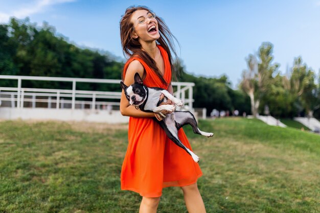 公園、夏のスタイル、陽気な気分で犬と遊ぶのを楽しんでオレンジ色のドレスで若い幸せな笑顔の女性