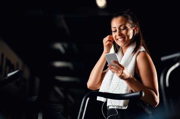 フィットネスセンターでワークアウトしながら携帯電話を使用して音楽を聴く若い幸せなスポーツウーマンコピースペース