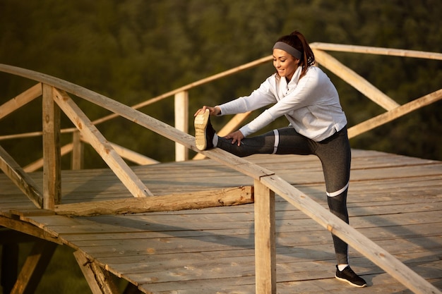 Young happy sportswoman warming up and stretching her legs on wooden bridge