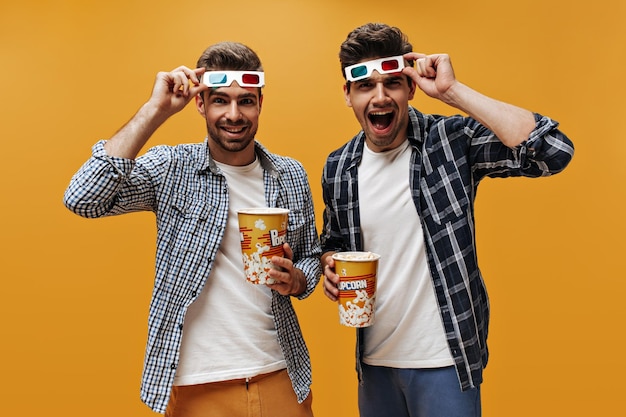 흰색 티셔츠와 파란색 체크무늬 셔츠를 입은 젊고 행복한 브루넷 남성들은 3D 안경을 벗고 영화를 보고 고립된 주황색 배경에 팝콘을 들고 있다