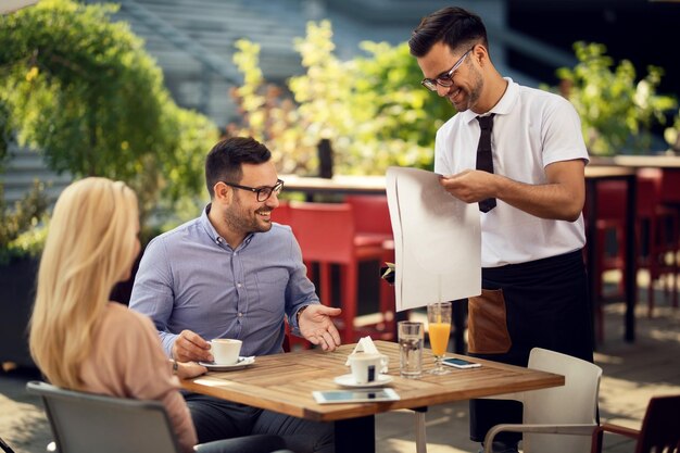 カップルにサービスを提供し、レストランで昼食のために彼らのテーブルを準備している若い幸せなウェイター