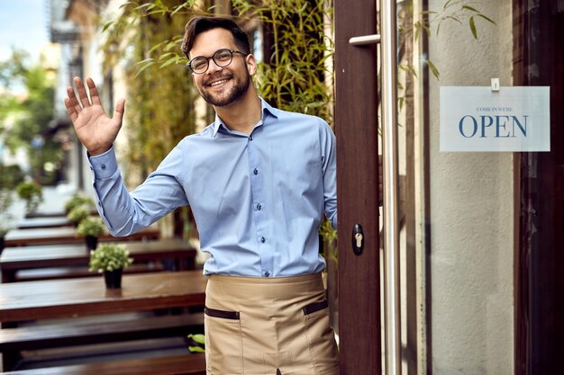 Young happy waiter standing at cafe's front door and waving to someone