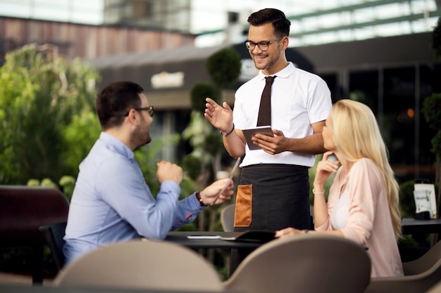 Free photo young happy waiter using digital tablet while talking to a couple and taking their order in a cafe