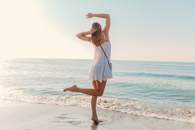 Молодая счастливая женщина танцует, поворачиваясь на берегу моря в солнечном летнем стиле моды в белом платье на каникулах