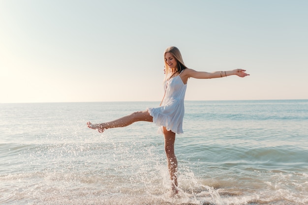 Молодая счастливая женщина танцует, поворачиваясь на берегу моря в солнечном летнем стиле моды в белом платье на каникулах